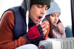 A man and a woman, bundled up in winter clothing including hats, gloves, and scarves, sit next to a radiator. The man warms his hands by blowing on them, while the woman holds her hands close to the heat source, appearing cold.