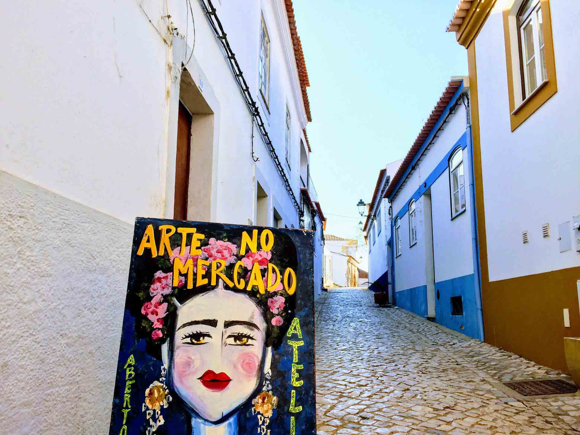 A narrow cobblestone street lined with white buildings in a sunny town. An art sign featuring a colorful woman’s face with flowers in her hair stands at the forefront. The sign reads "Arte no Mercado." The blue sky is clear above the warm, inviting street scene.