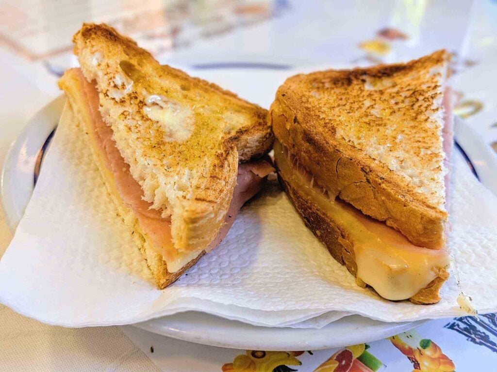 A close-up image of a grilled cheese sandwich cut diagonally and placed on a white plate with a napkin. The sandwich features golden-brown toasted bread with melted cheese oozing out from inside. The plate, reminiscent of Portugal's charming cafes, is set on a table with a floral patterned tablecloth.