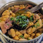A metal serving dish filled with a hearty Portuguese stew of white beans, chunks of meat, and garnished with chopped fresh parsley. A spoon rests inside the dish, and it sits on a blue and white patterned plate.
