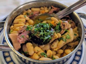 A metal serving dish filled with a hearty Portuguese stew of white beans, chunks of meat, and garnished with chopped fresh parsley. A spoon rests inside the dish, and it sits on a blue and white patterned plate.