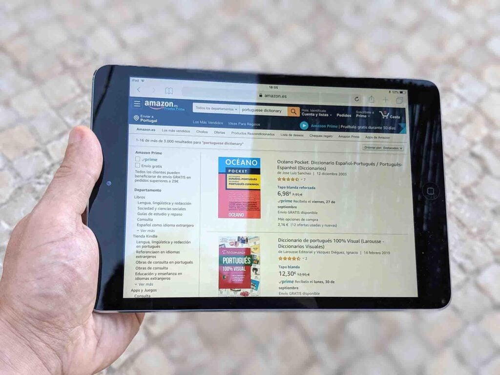 A person is holding a tablet displaying an Amazon webpage. The webpage shows a search for Portuguese dictionaries, featuring listings for "Oceano Pocket Dictionary" and "Dicionário de Portugais 100% Visual." The handheld tablet has a black case.