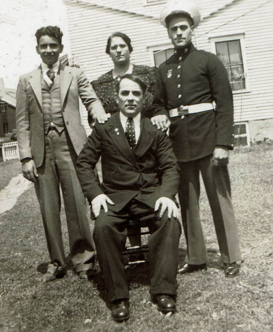 A vintage black-and-white photo of four individuals. Three men and one woman pose outside in front of a house. One man sits in a chair wearing a suit, while the two other men, one in a suit and one in a military uniform, and the woman stand behind him.