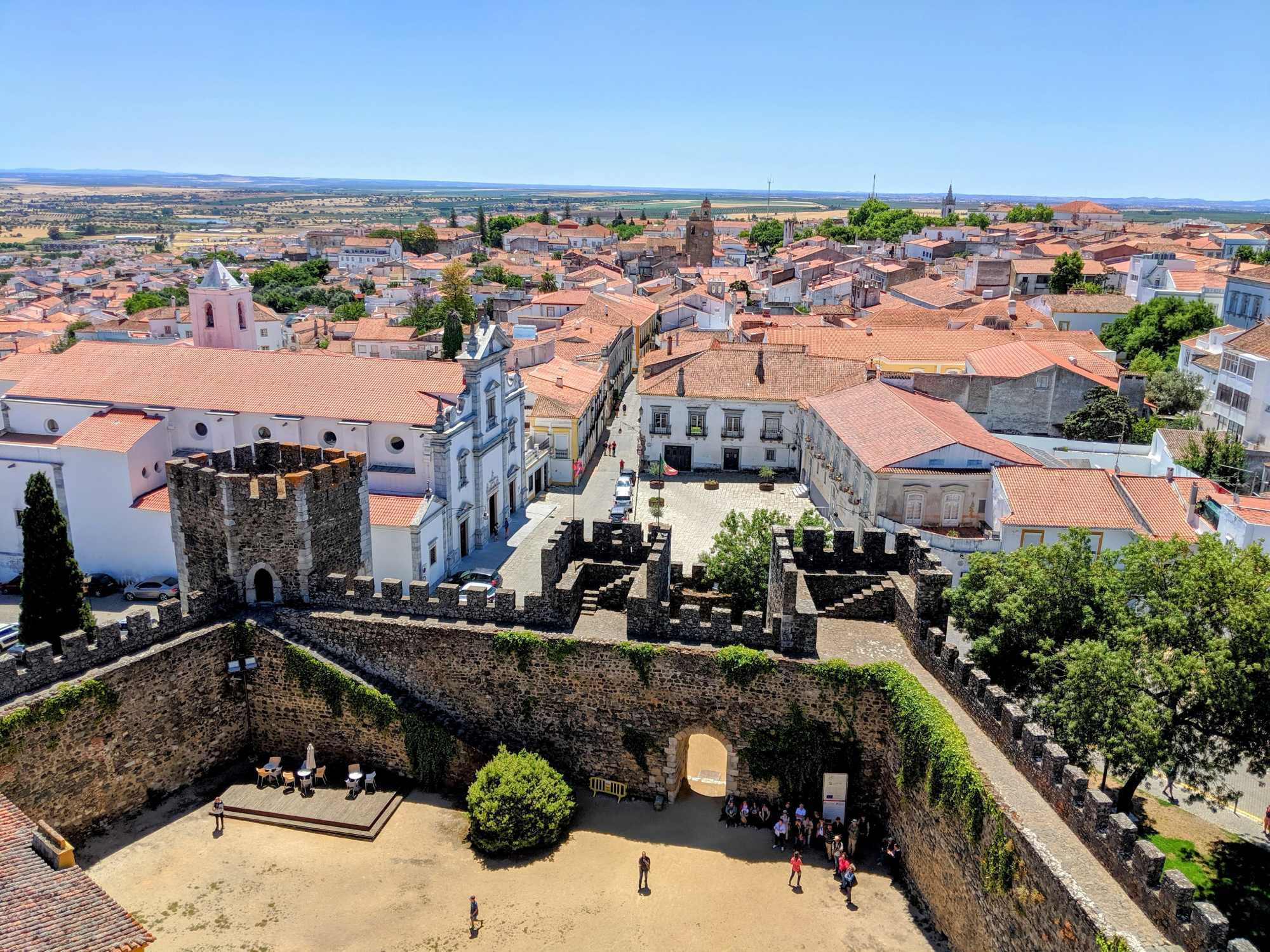 Aerial view of Castelo de Portalegre in Portugal, showcasing its stone walls and courtyard with people gathered inside. Beyond the castle, you can see the town's red-roofed buildings and the picturesque landscape of Portugal under a clear blue sky.