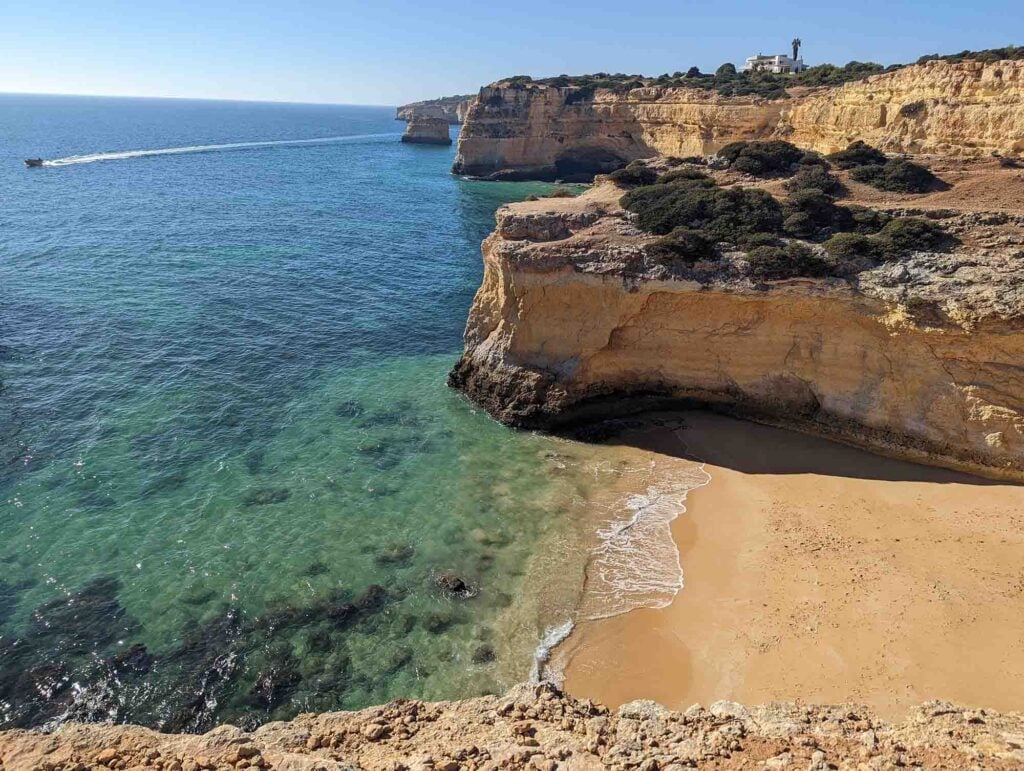 A secluded beach in the Algarve sits at the base of tall, rugged cliffs. The clear turquoise water contrasts with the golden sand and rocky cliffs, stretching out to the horizon. A boat can be seen traveling on the calm ocean, leaving a white trail behind it.