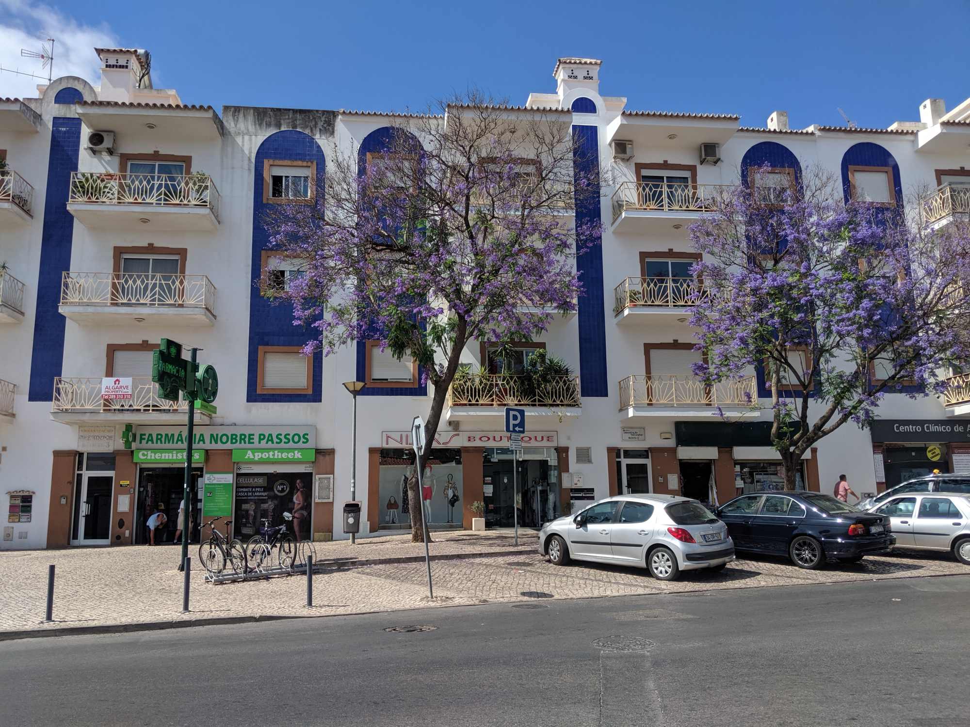A street view of a white building with blue accents in Portugal, featuring three stories with balconies. Cars are parked in front, and a pharmacy, boutique, and other shops occupy the ground level. Blooming purple trees line the sidewalk under a bright sky.