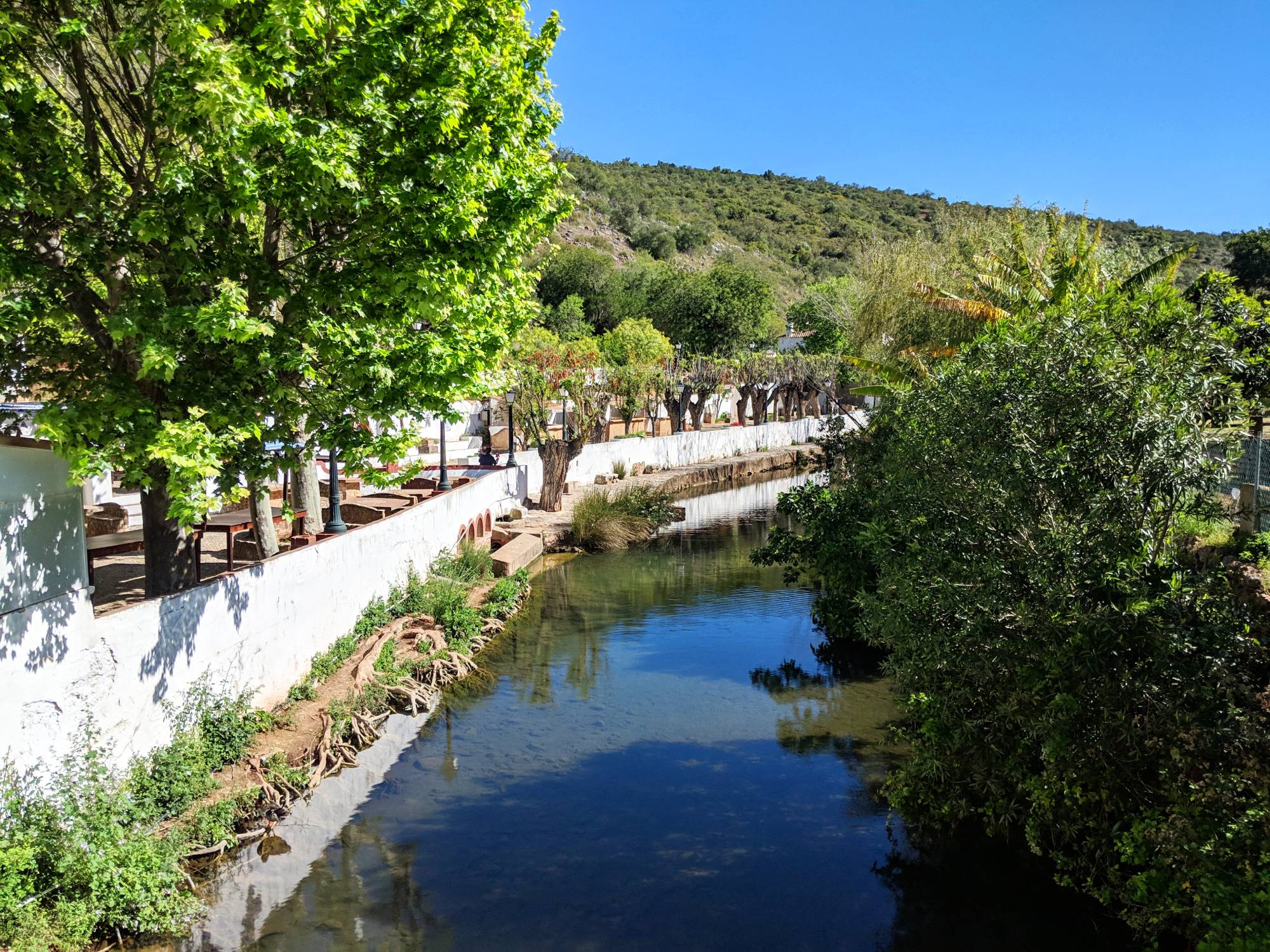 A serene stream flows through a lush, green landscape in Portugal, with trees on either side. A white wall lines the left bank, and there are outdoor seating areas visible behind it. The clear blue sky above and the tranquil water below reflect the greenery around.