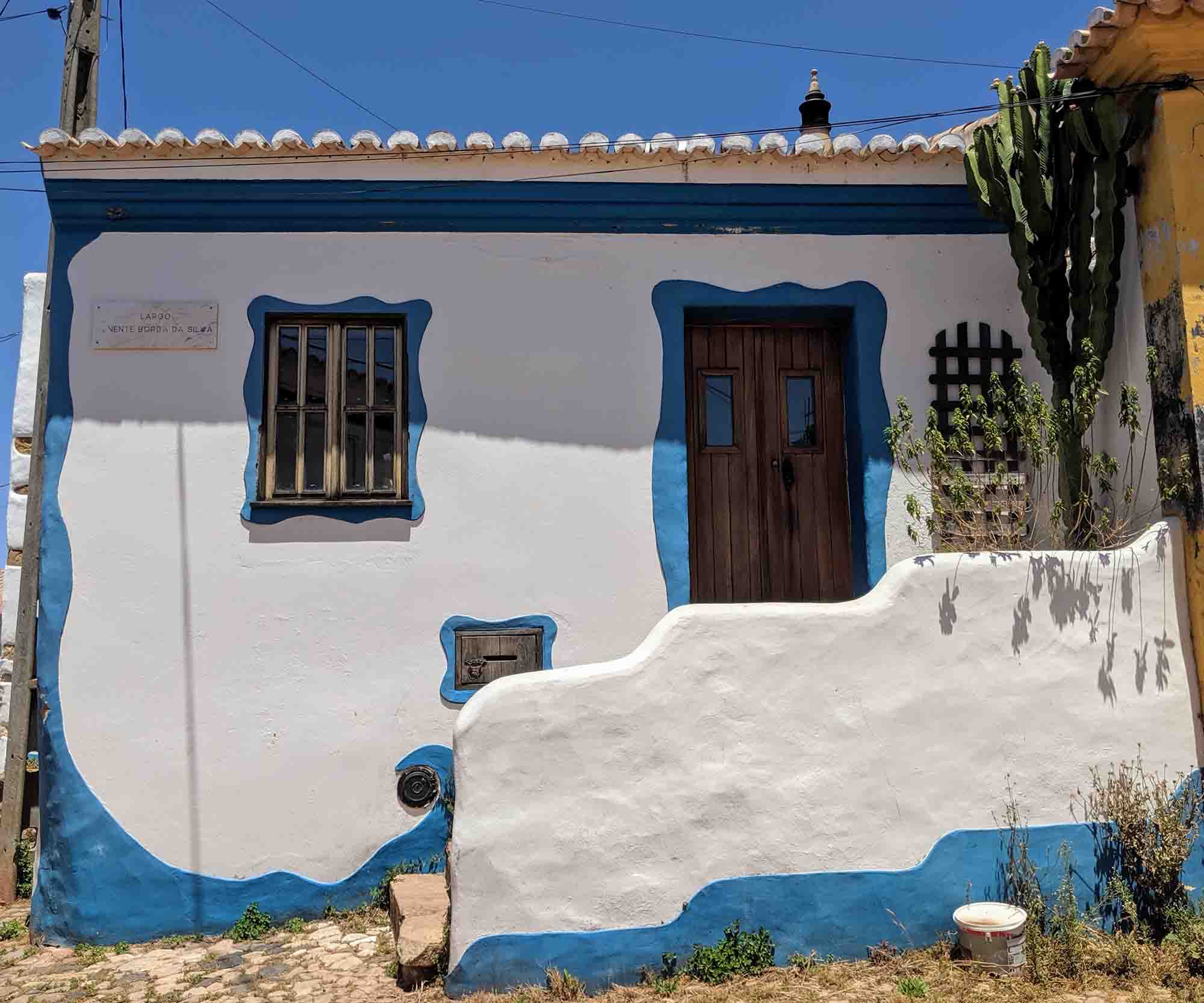 A small white house with a wavy blue trim around the door, window, and lower wall. The wooden door and window have bars, and a cactus grows on the right side. A mosaic tile nameplate is on the upper left. The house, reminiscent of Portugal's charm, is situated under a clear blue sky.