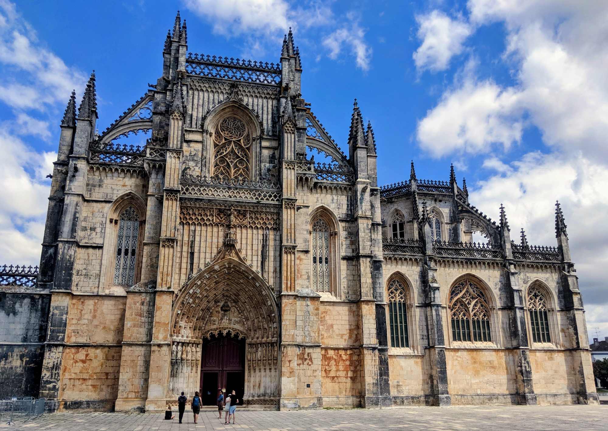 A grand, Gothic-style cathedral in Portugal with intricate stone carvings and tall spires against a blue sky with clouds. The facade features large arched windows and a detailed entrance. Several people are standing in front of the building, emphasizing its impressive scale.