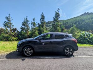 A black SUV is parked on a roadside in the Azores. The background features vibrant greenery, tall trees, and hills under a clear blue sky. The sun casts bright light, highlighting the side profile of the vehicle and the serene, lush landscape.