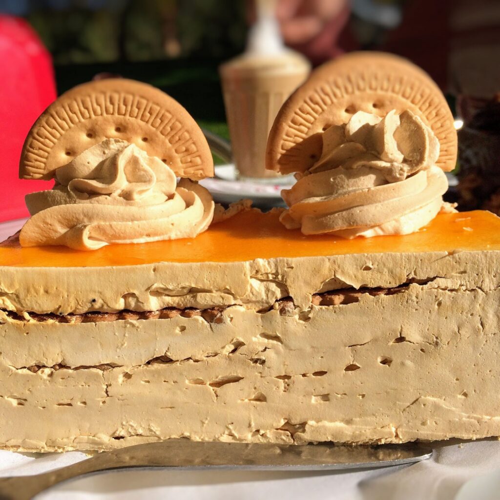 A rectangular layered bolo de bolacha cake with a smooth beige exterior is topped with two dollops of swirled light brown frosting. Each dollop is adorned with a half-moon-shaped cookie, reminiscent of desserts enjoyed in Portugal. The cake sits on a white surface, with a blurred background of an outdoor setting.