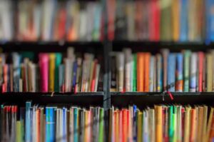 A blurred image of a bookshelf filled with various colorful books. The books are arranged in multiple rows, and the focus is soft, making it difficult to distinguish individual titles or authors. The shelves appear to be packed with a variety of books.