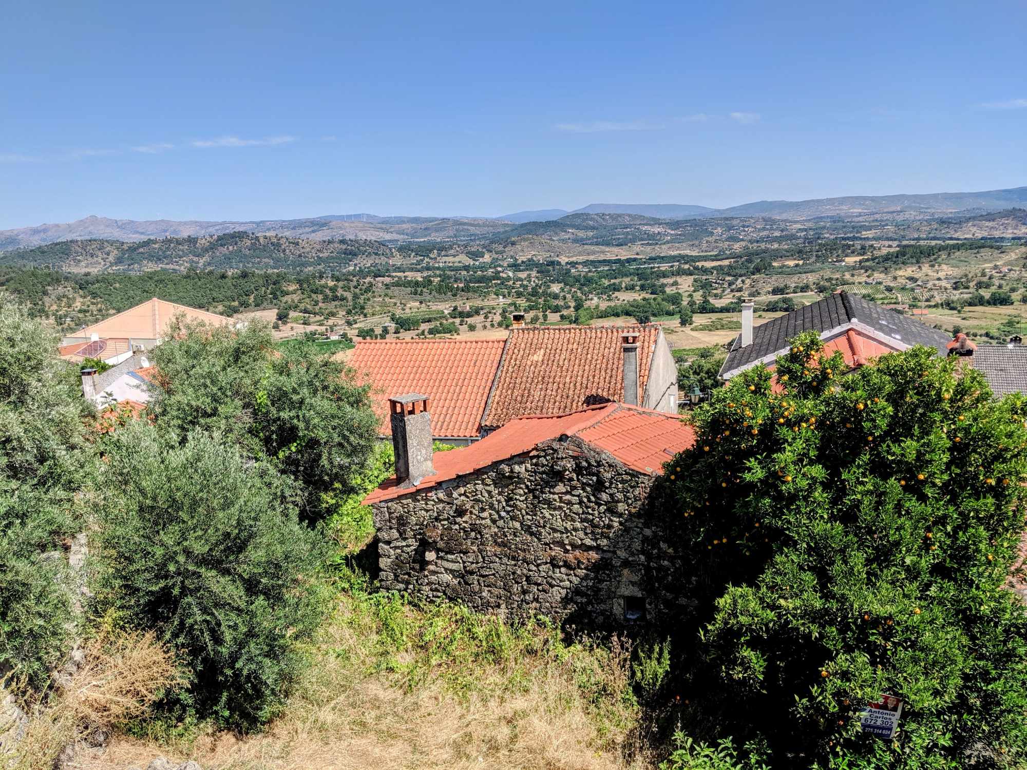 Scenic landscape of a rural area in Portugal with red-tiled rooftops of stone houses, greenery, and trees in the foreground. The backdrop features rolling hills and mountains under a clear blue sky.