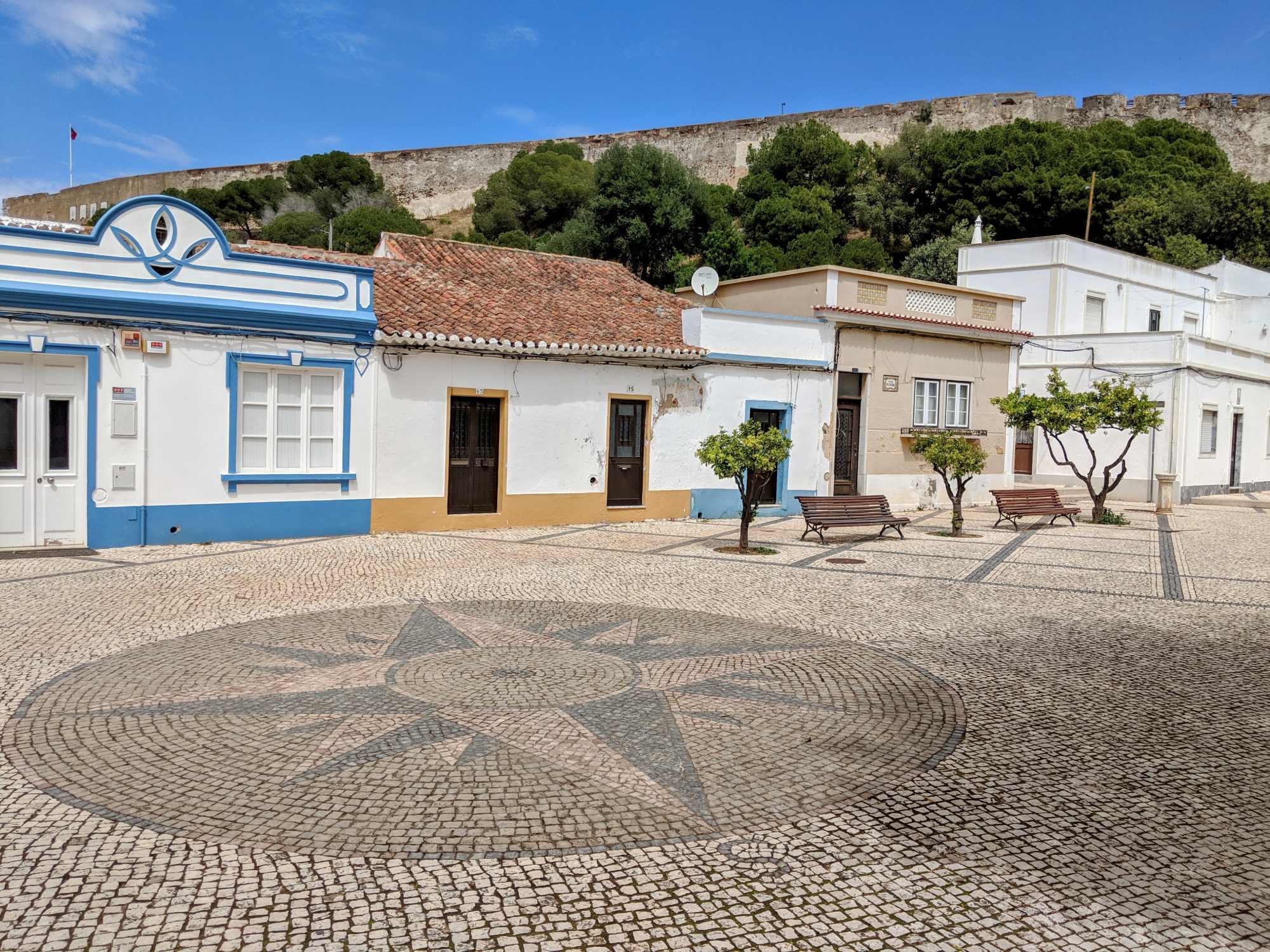 A sunlit town square with patterned cobblestone pavement featuring a circular mosaic design. Surrounding buildings are white with blue accents, reminiscent of Portugal's charming architecture. A hill with a fort atop and green trees are in the background. Benches and small trees line the square.