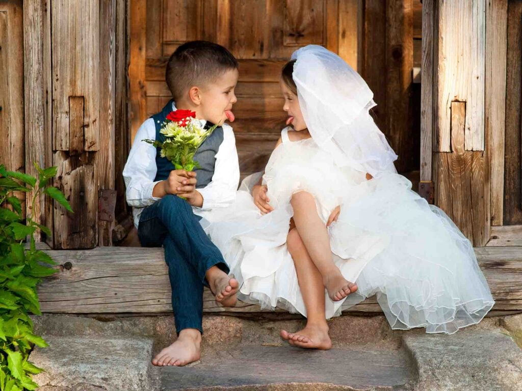 Two barefoot children dressed as bride and groom sit on wooden steps, playfully interacting—likely at a celebration in Portugal.