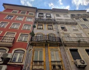 A view looking up at four adjacent old buildings with colorful, intricate facades. The left building is red, the center-left is yellow, the center-right is white, and the right building is beige. Each building has balconies and multiple windows, with a blue sky above.
