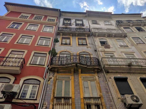 A view looking up at four adjacent old buildings with colorful, intricate facades. The left building is red, the center-left is yellow, the center-right is white, and the right building is beige. Each building has balconies and multiple windows, with a blue sky above.