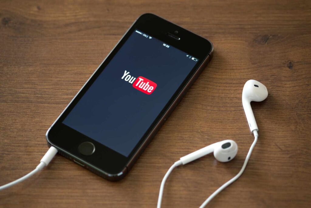 A smartphone on a wooden surface displays the YouTube logo on its screen. A pair of white earbuds is connected to the phone, with one earbud positioned near the phone and the other lying slightly further away.