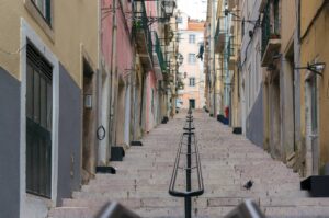A narrow, steep staircase in Alfama lined with colorful buildings on both sides in an urban setting. The left side features pastel yellow and pink walls, while the right side has earthy tones. A pigeon is walking near the bottom, and a person is visible in the distance.