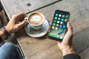 A person is holding a smartphone in their right hand, displaying the home screen with various app icons. Their left hand is holding a white cup of coffee with foam on top. The items are on a wooden table, and the person is wearing a watch on their left wrist.