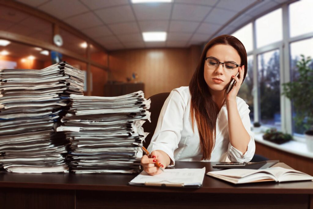 A woman on the phone, sitting next to two piles of backlogged files