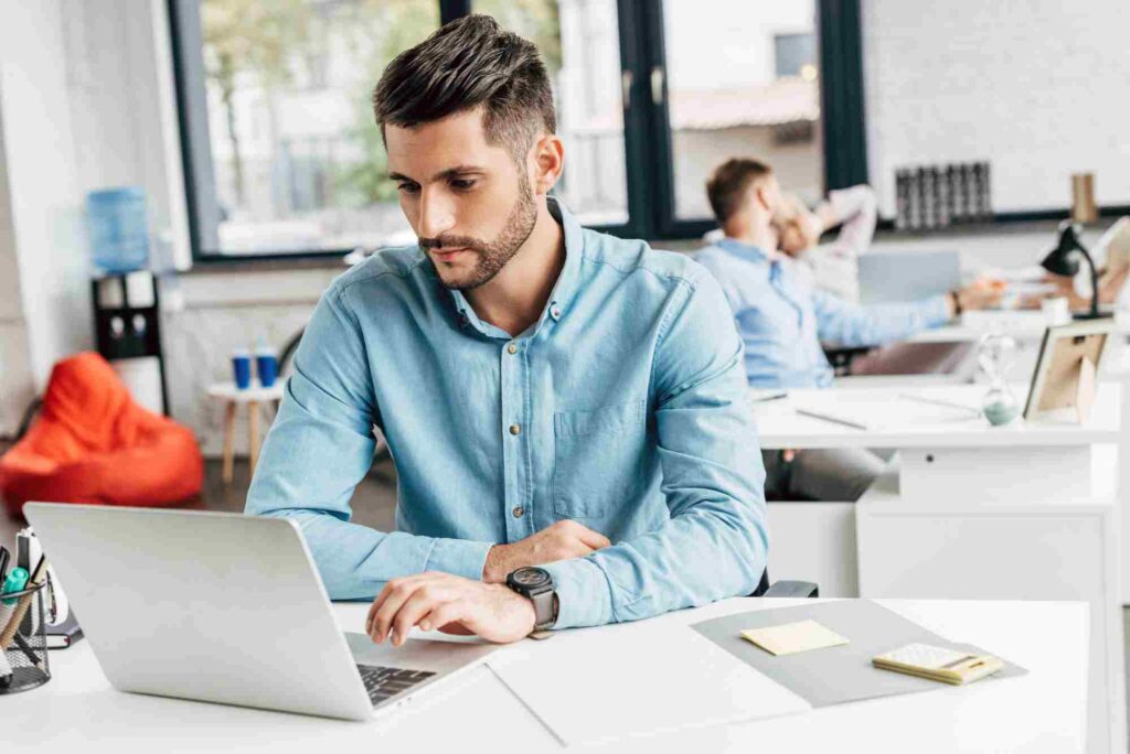 A man with dark hair and a beard is working on a laptop at a desk in a modern, open-plan office. He is wearing a blue shirt and a black smartwatch. In the background, another person is talking on the phone at a different desk.