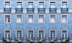 A symmetrical facade of a building covered in blue and white tiles with 15 windows in a grid pattern. Each window has a blue wrought-iron balcony railing. The design is consistent across the entire surface, creating a harmonious and repetitive visual effect.