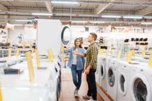 A man and a woman are in an appliance store. They stand together examining a front-loading washing machine. The store is filled with various household appliances, including washing machines and other equipment displayed on shelves.