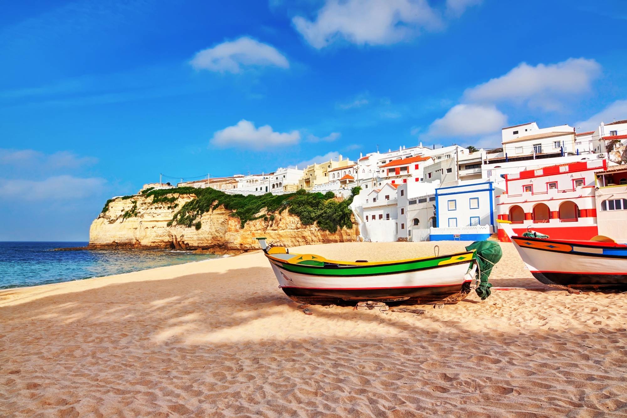 A picturesque beach scene in Carvoeiro features colorful fishing boats resting on the sandy shore. In the background, whitewashed buildings with vibrant accents are perched on a cliffside. The blue sky is dotted with a few clouds, complementing the tranquil setting.