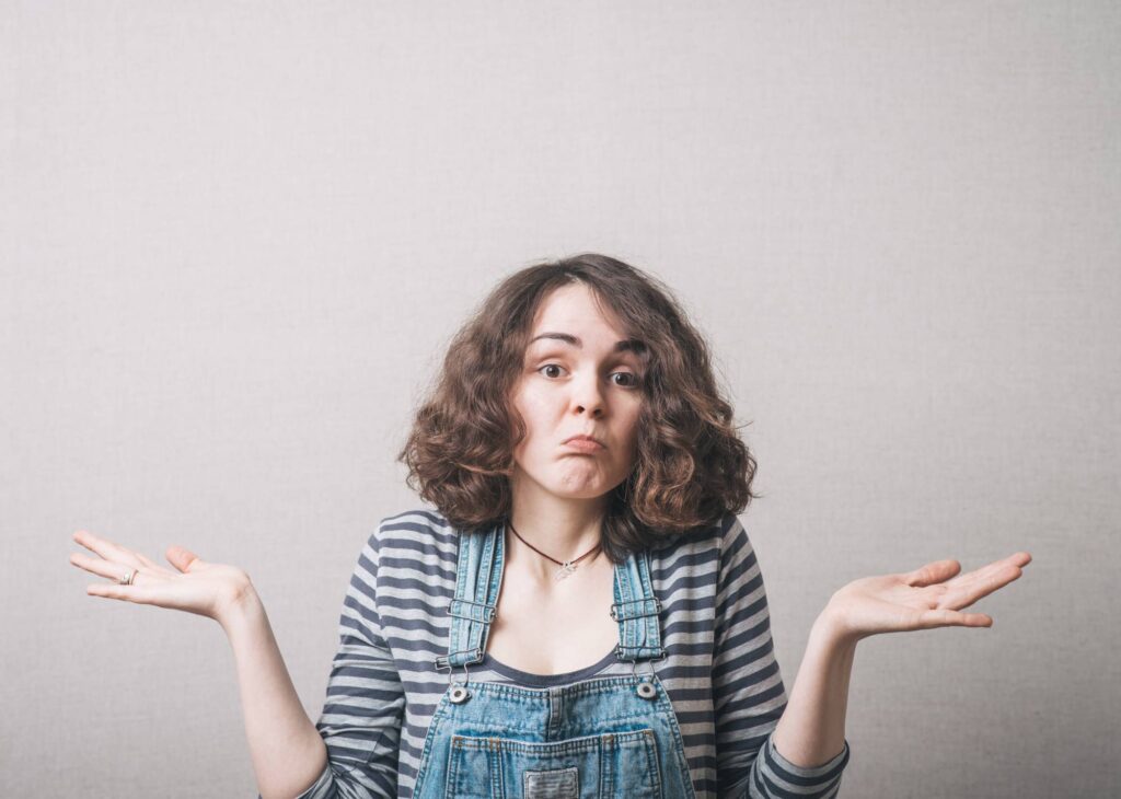 A person with shoulder-length curly hair wearing a striped shirt and denim overalls is standing against a plain background with their arms outstretched and palms up, displaying a puzzled or unsure expression.