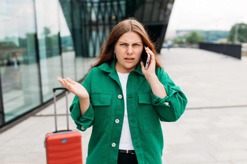 A woman with long brown hair in a green jacket stands outside with a red suitcase, looking confused while calling, possibly about traveling to Portugal.