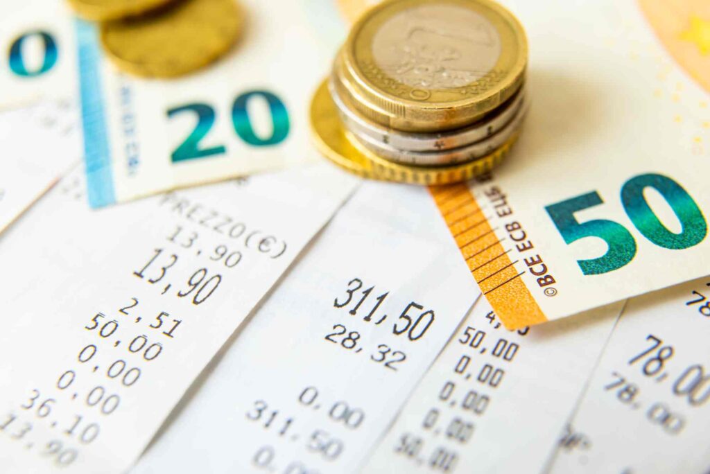 Close-up image showing receipts, Euro banknotes, and several stacks of Euro coins. The receipts display various purchase amounts, while the banknotes include denominations of 20 and 50 Euros. Coins are shown in different sizes and stacked on the receipts and banknotes.