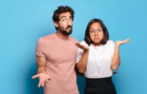 A man and woman stand against a blue background, shrugging with puzzled faces, suggesting uncertainty about moving to Portugal.
