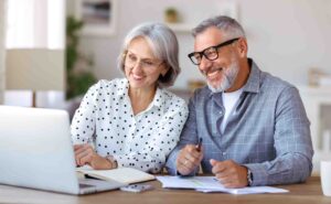 An older couple sits at a table, smiling and looking at a laptop screen. Both wear glasses; the woman is in a white polka-dot blouse, and the man is in a grey checked shirt. They seem to be working together, with papers and a notebook on the table.