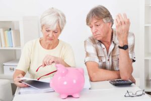 An older woman and man sitting at a table looking concerned. The woman is holding paperwork and red glasses, while the man has his hand raised and is looking at a calculator. A pink piggy bank is placed in front of them on the table.
