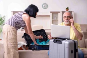 An older man sits on a couch, using a laptop with a phone to his ear, while a woman stands nearby packing a suitcase. The room is bright, with light-colored furniture and a clock on the wall behind them.