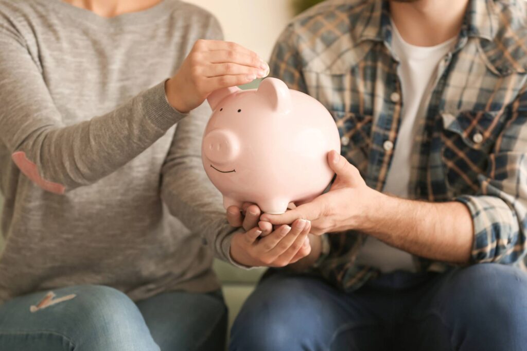 Two people are seen from the shoulders down, placing a coin into a pink piggy bank. The woman on the left, in a gray sweater, is dropping the coin while the man on the right, in a plaid shirt, is holding the piggy bank.