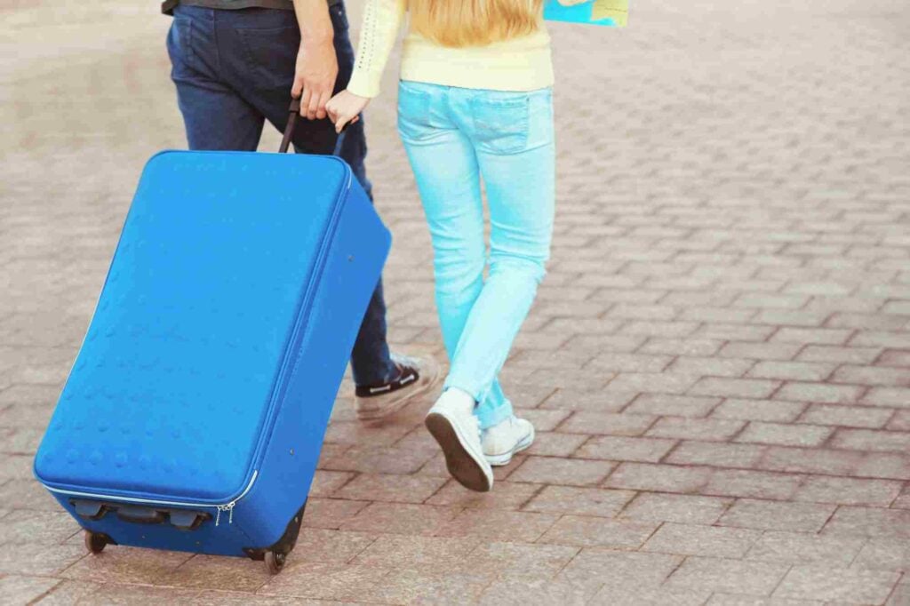 A man and a woman are walking on a paved path, holding hands. The man is pulling a large blue suitcase on wheels with his other hand. The couple's faces are not visible, and they appear to be traveling or on a journey.