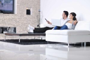 A couple sits on a white couch in a modern living room, watching TV. One holds a remote control, and the other has a laptop. The room features a stone accent wall, sleek furniture, and a black rug on a polished floor.