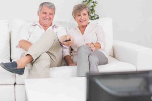 A smiling elderly couple sits on a white couch in a bright living room, both looking towards a television. The man has his legs crossed and the woman is holding a remote control. They appear relaxed and are leaning slightly towards each other.