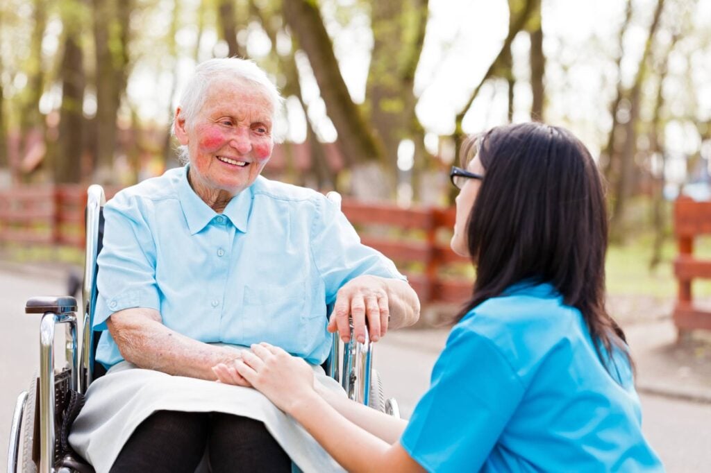 An elderly woman in a wheelchair holds hands with her caregiver outdoors in Portugal, both smiling on a sunny day near trees and a fence.