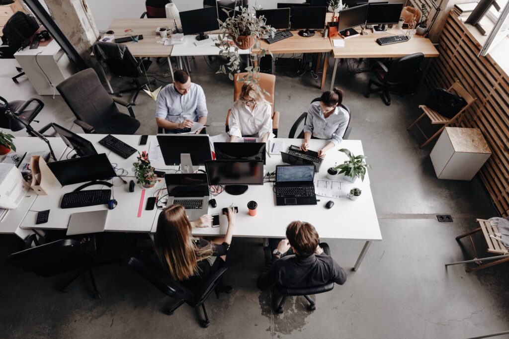 A modern office space with five people working at their desks. The desks have multiple computer monitors, laptops, and office supplies. The office has a bright, open layout with large windows, plants, and a mix of wooden and white furniture.