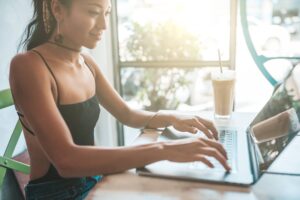 A woman sits at a table in a bright, sunlit cafe, working on a laptop. She wears a black tank top and a choker necklace. Beside her is a tall glass of iced coffee with a straw. The background shows large windows with blurred greenery outside.