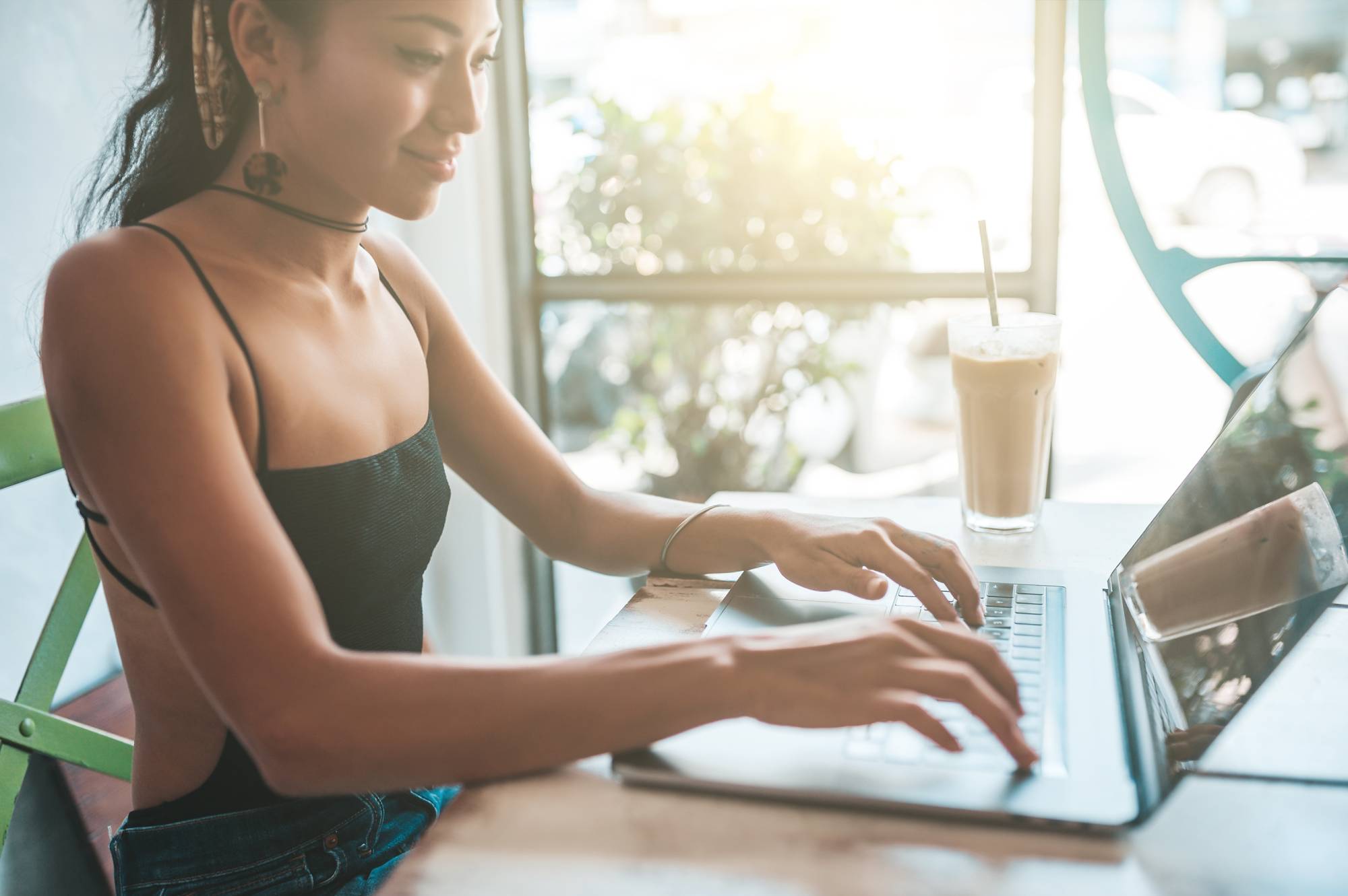 A woman sits at a table in a bright, sunlit cafe, working on a laptop. She wears a black tank top and a choker necklace. Beside her is a tall glass of iced coffee with a straw. The background shows large windows with blurred greenery outside.