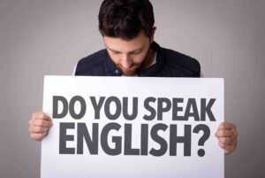 A man with a beard is looking down while holding a large white sign that reads "DO YOU SPEAK ENGLISH?" in bold, black letters. He is wearing a black vest over a gray shirt and standing against a plain, gray background.