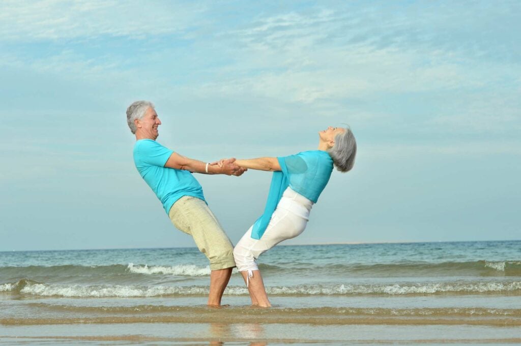 An elderly couple stands in shallow ocean water, holding hands and leaning back, with smiles on their faces. Both wear light blue tops and light-colored pants, enjoying a playful moment on a sunny day at the beach. The sky is clear with a few clouds.