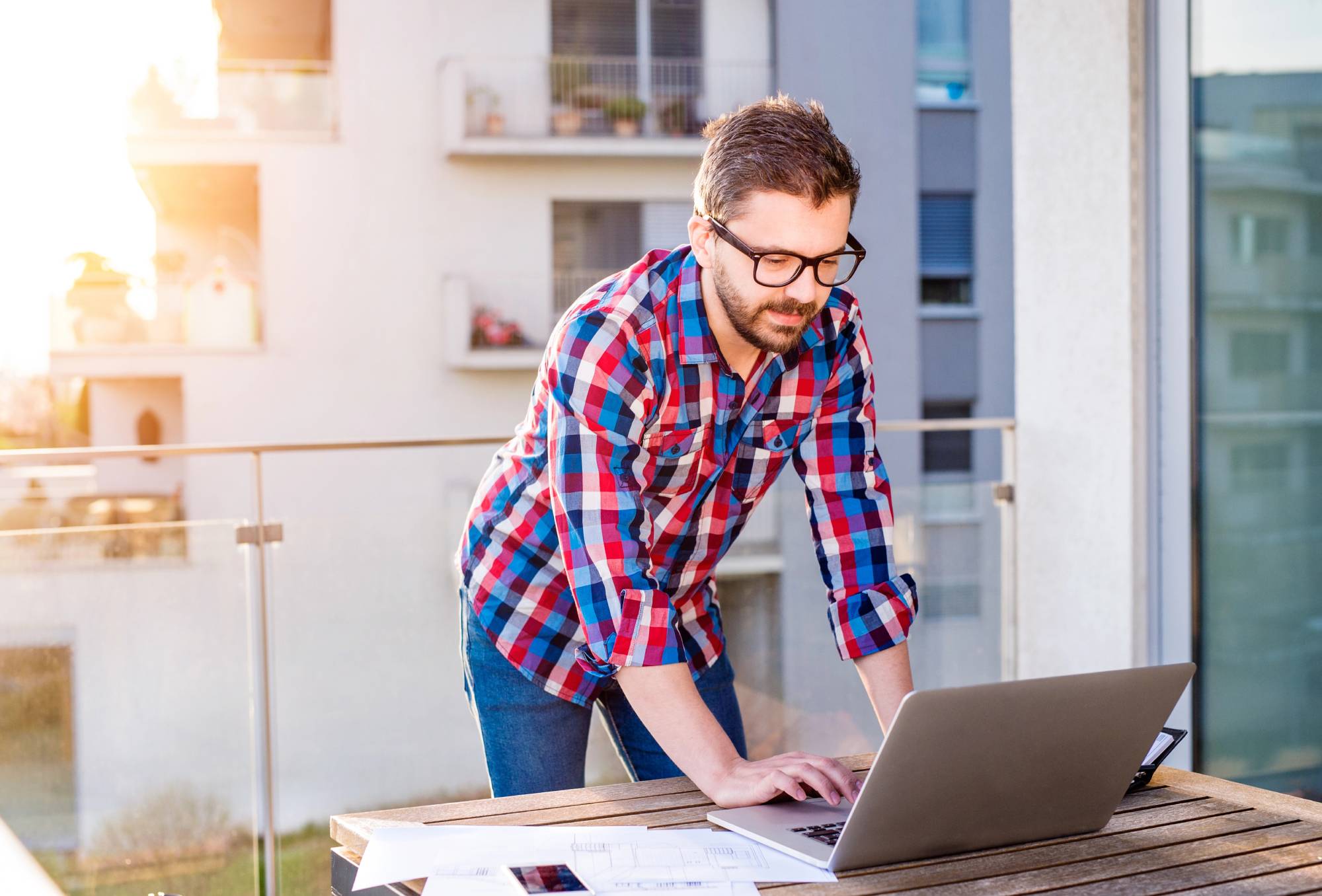A man with a beard and glasses is standing on a balcony, leaning over a wooden table while working on a laptop. He is wearing a red and blue checkered shirt. The background shows a building in daylight.