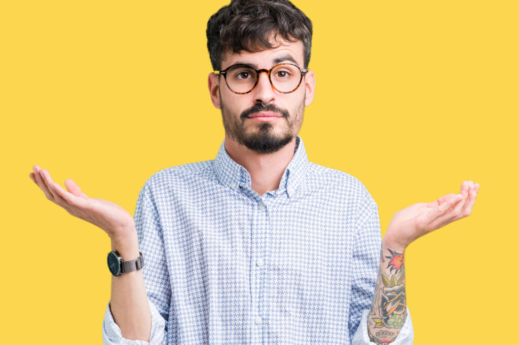 A young man with glasses and a beard shrugs, looking uncertain. He has tattoos on one arm, set against a yellow background.