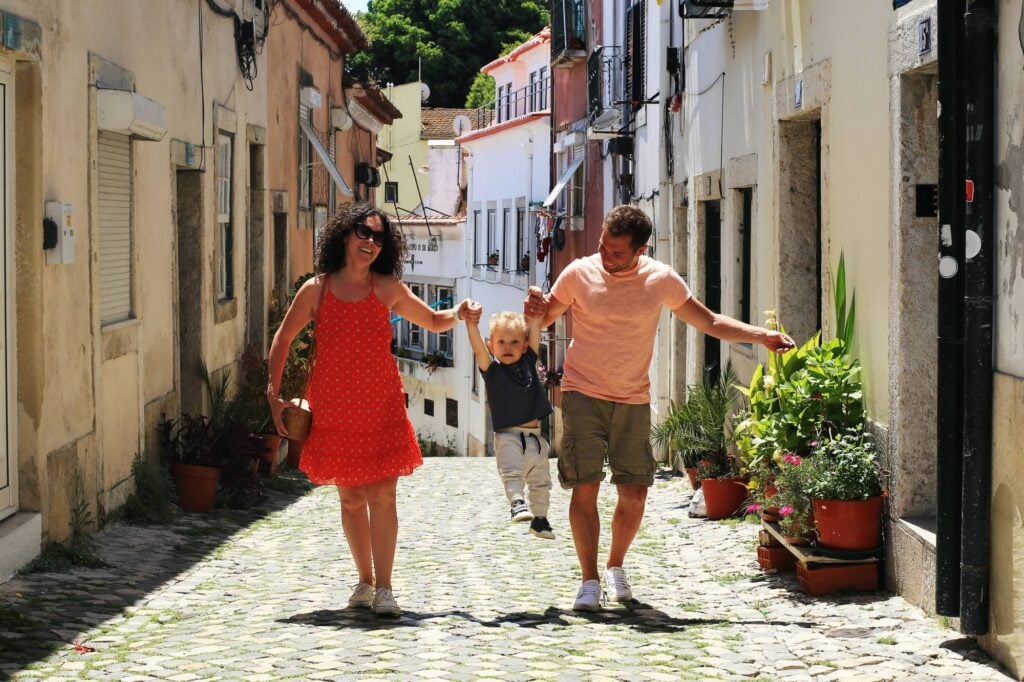 A happy family swings a child between them while strolling down a sunlit, old Portuguese cobblestone street in Lisbon with plants and historic buildings.