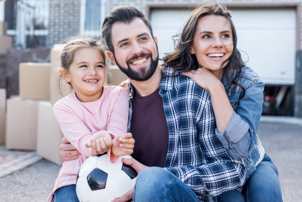 A smiling family of three poses outside, sitting close together. A young girl in a pink shirt holds a soccer ball, with her father in a plaid shirt kneeling beside her and her mother in a denim jacket standing behind, embracing them.